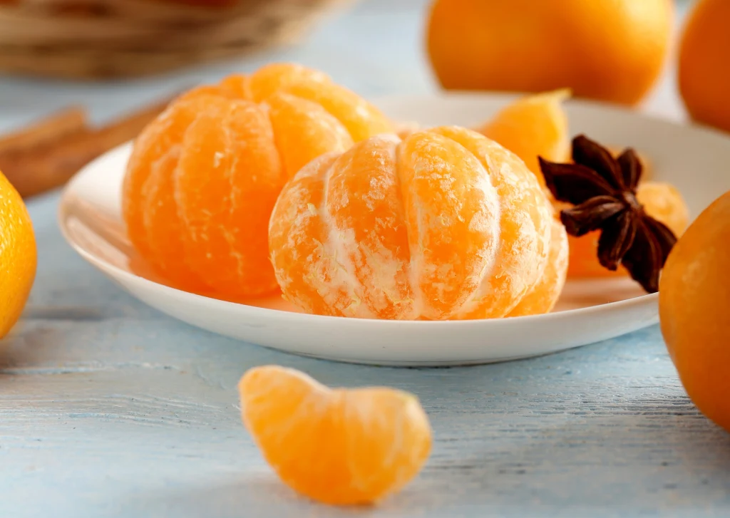 Obrane mandarynki ułożone na białym talerzu obok kilku cząstek owocu i anyżu gwiaździstego, w tle widoczne całe mandarynki i delikatnie rozmyty kosz.