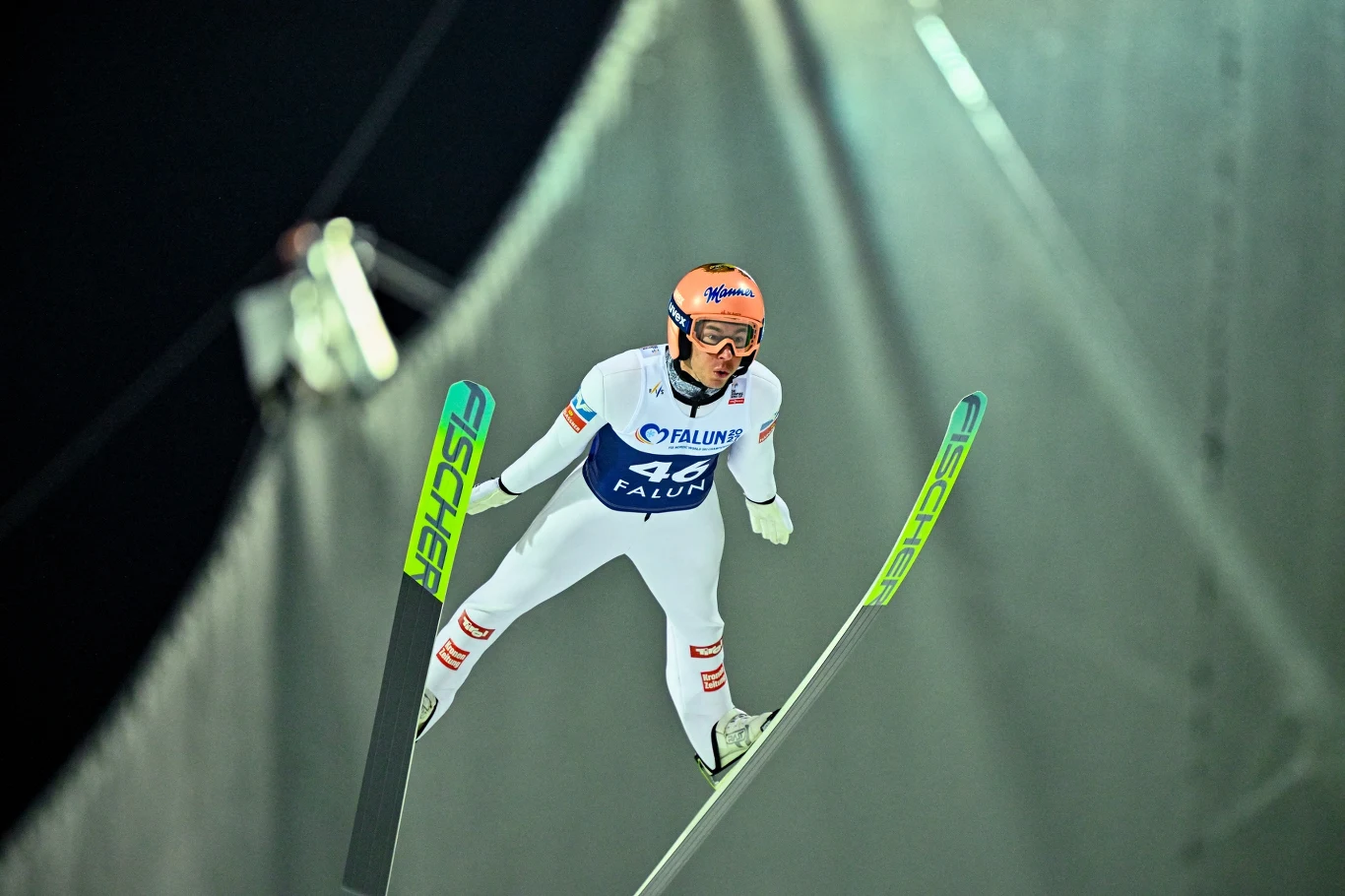 Zawodnik w kombinezonie narciarskim szybujący w powietrzu podczas skoku narciarskiego. Sportowiec ma na głowie kask i gogle ochronne, obie narty są skierowane równolegle, a w tle widoczna jest rampa skoczni.