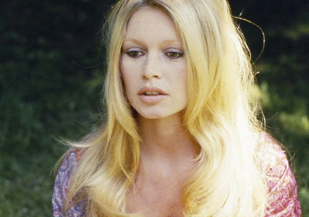 Blondynka o długich włosach ubrana w fioletowo-różową bluzkę siedzi na tle zieleni, jej twarz jest lekko zamyślona, a spojrzenie skierowane w dół.