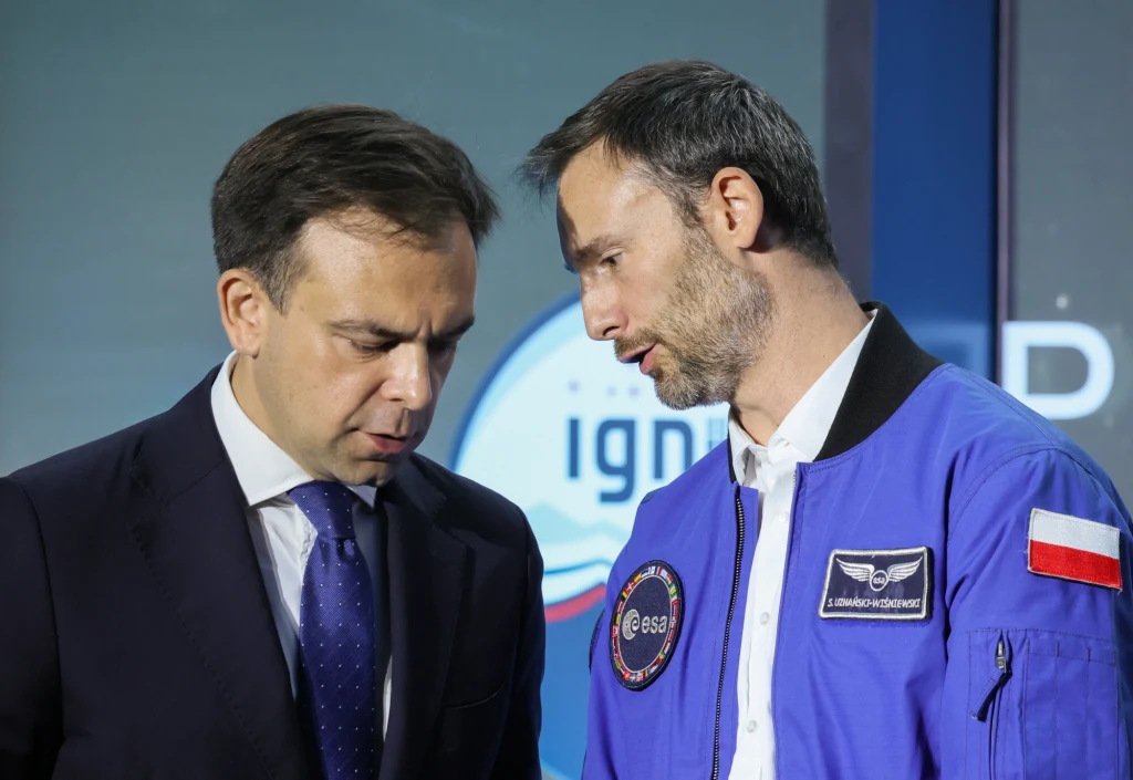 Dwóch mężczyzn podczas konwersacji, z których jeden ubrany jest w garnitur, a drugi w niebieski kombinezon z emblematami ESA oraz polską flagą na ramieniu. W tle widoczne logo związane z tematyką kosmiczną.