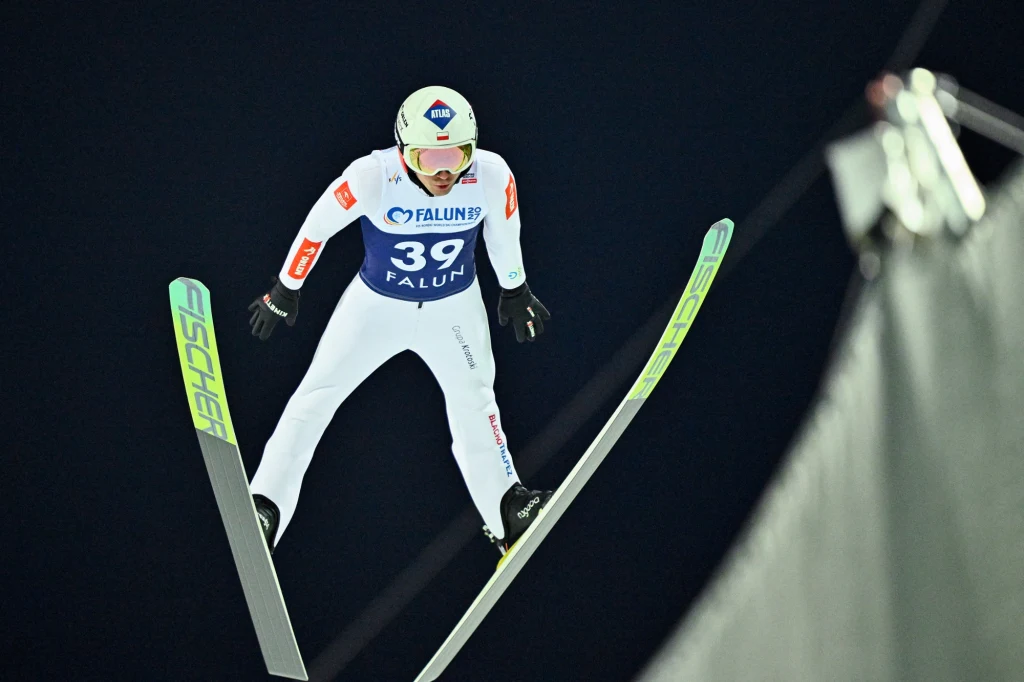 Kamil Stoch Zawodnik w białym kombinezonie startowym z numerem 39 wykonuje skok narciarski na tle ciemnego nieba podczas zawodów w Falun, z widocznymi nartami typu Fischer.