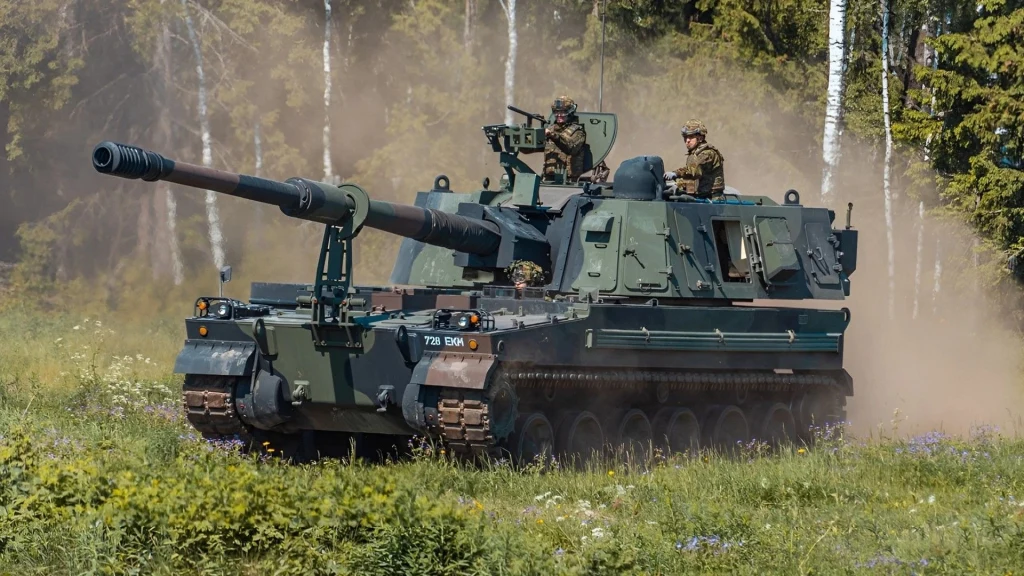 Estonia obawia się inwazji Rosji. Zakupiła ogromną ilość broni z USA Samobieżna armatohaubica w trakcie przemieszczania się przez zalesiony teren, dwóch żołnierzy na pokładzie, unoszący się kurz wokół pojazdu, w tle drzewa i zieleń.