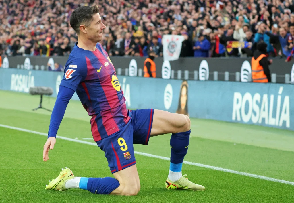 Robert Lewandowski Piłkarz w stroju FC Barcelony świętuje strzeloną bramkę wykonując efektowny ślizg na kolanach na murawie, w tle kibice na trybunach z transparentami i flagami.