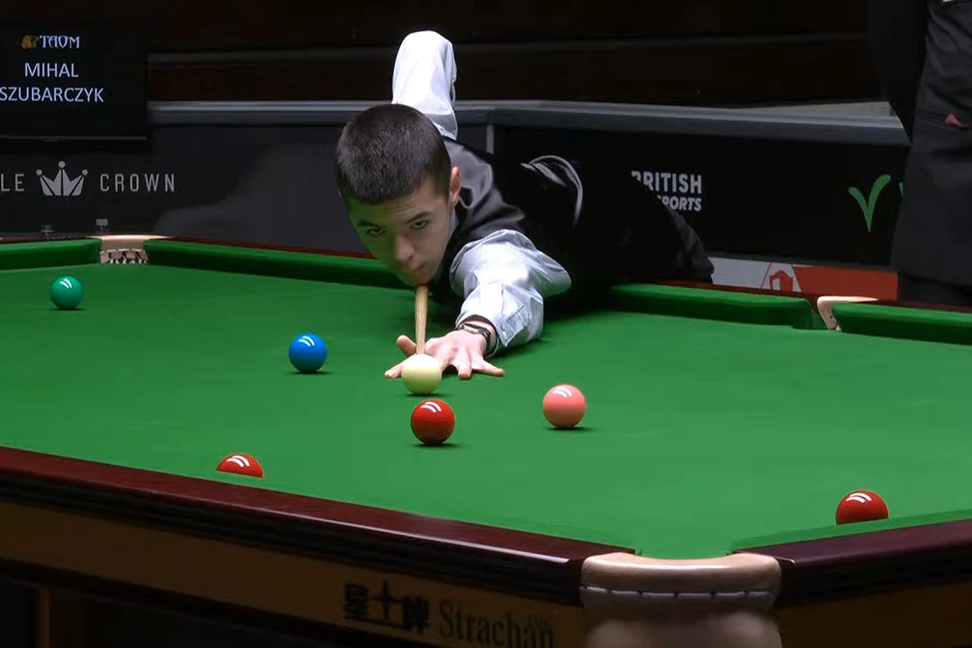 Młody zawodnik skupiony na wykonaniu precyzyjnego zagrania przy stole do snookera, wokół niego rozmieszczone kolorowe bile, w tle fragment widowni i tablica z nazwiskiem gracza.