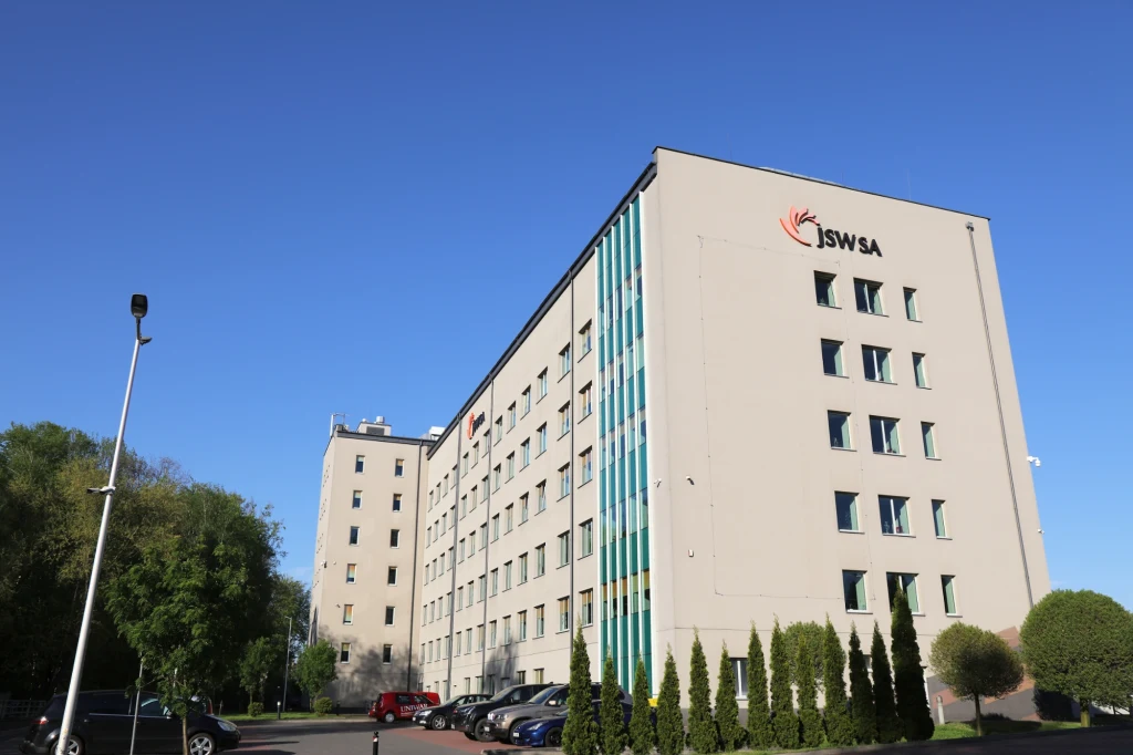 Problemy finansowe Jastrzębskiej Spółki Węglowej narastają Nowoczesny, wielokondygnacyjny budynek o jasnej elewacji z logotypem JSW SA, otoczony zielenią, z zaparkowanymi samochodami na parkingu oraz lampami ulicznymi, po lewej stronie drzewa i krzewy, niebo bezchmurne.