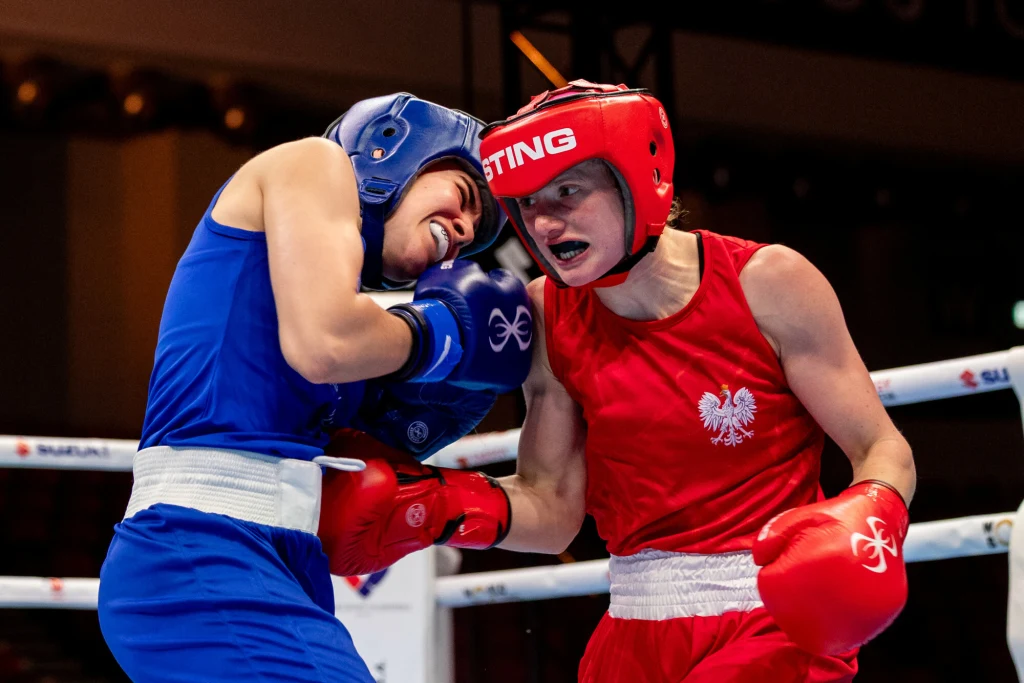 Natalia Kuczewska (w czerwonym stroju) wróci z Budapesztu z medalem ME U-23 Dwóch bokserów w trakcie walki w ringu, jeden ubrany w niebieski strój ochronny, drugi w czerwonym z orłem na piersi, zacięcie i determinacja widoczne na ich twarzach, otoczenie sportowej areny, jasne oświetlenie.