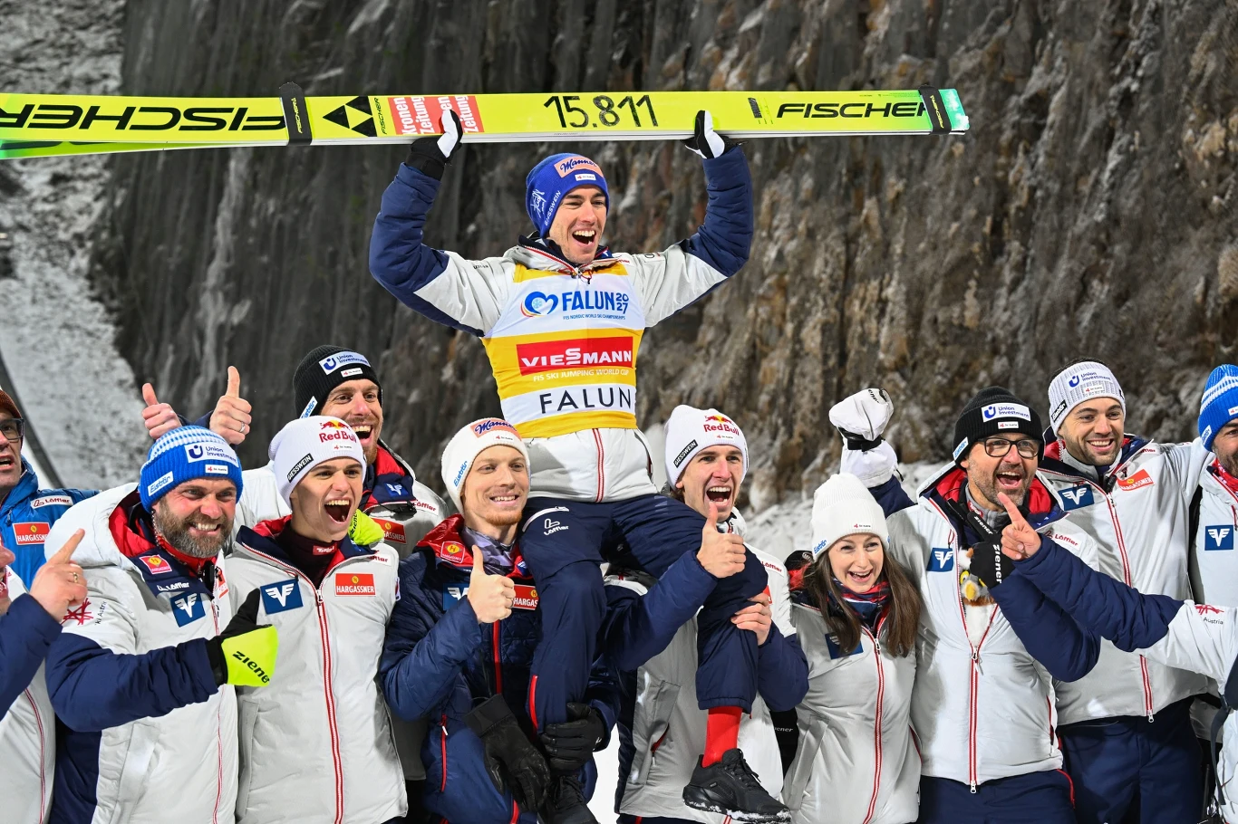 Zwycięski skoczek narciarski wznosi narty nad głowę w geście radości, siedząc na barkach kolegi z drużyny otoczony uśmiechniętym zespołem ubranym w biało-granatowe stroje, wszyscy świętują sukces na tle zimowego krajobrazu.