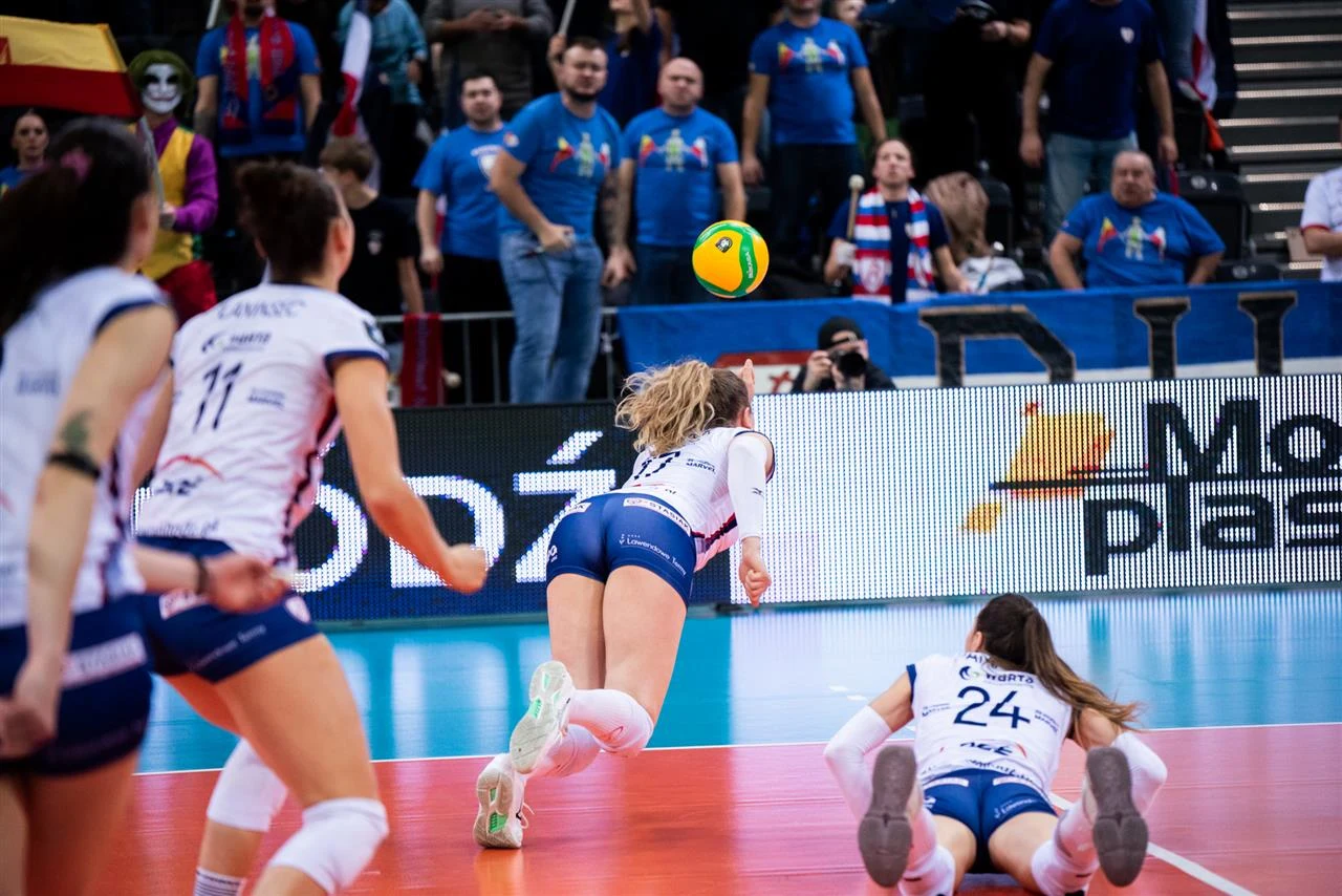 Zawodniczki drużyny siatkarskiej w dynamicznej akcji na parkiecie, jedna z nich rzuca się po piłkę, podczas gdy inne obserwują przebieg sytuacji. W tle widownia uważnie śledzi rozgrywkę.