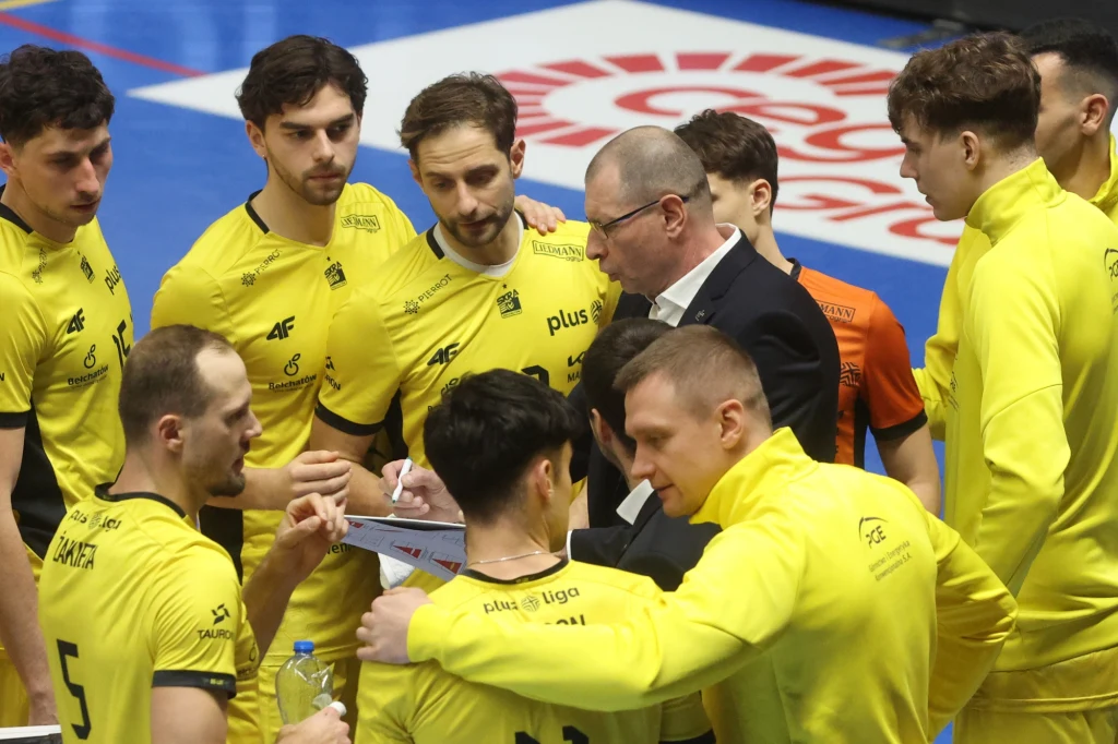 Grupa siatkarzy ubranych w żółte stroje wraz z trenerem omawia strategię podczas przerwy w meczu, wokół nich panuje skupienie i zaangażowanie.