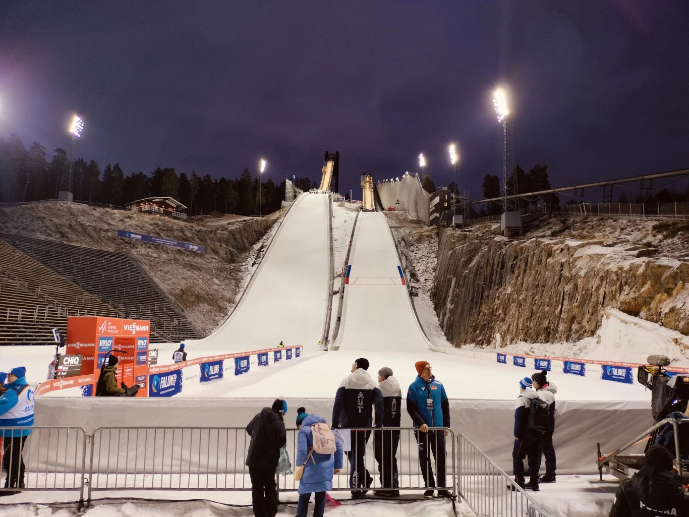 Oświetlona skocznia narciarska otoczona trybunami i barierkami ochronnymi, w dolnej części grupy ludzi w zimowych ubraniach, zorganizowana infrastruktura sportowa, widoczne reklamy sponsorów i sędziowie przygotowujący wydarzenie.