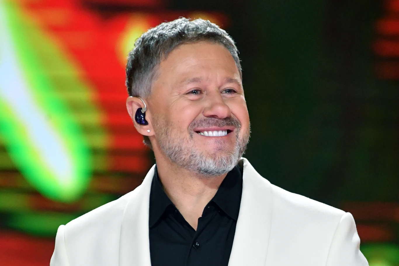 Andrzej Piaseczny nagrał z grupą BEMY swój przebój po francusku Uśmiechnięty mężczyzna w białej marynarce i czarnej koszuli, na tle rozmytego, kolorowego tła, z widoczną słuchawką w uchu.