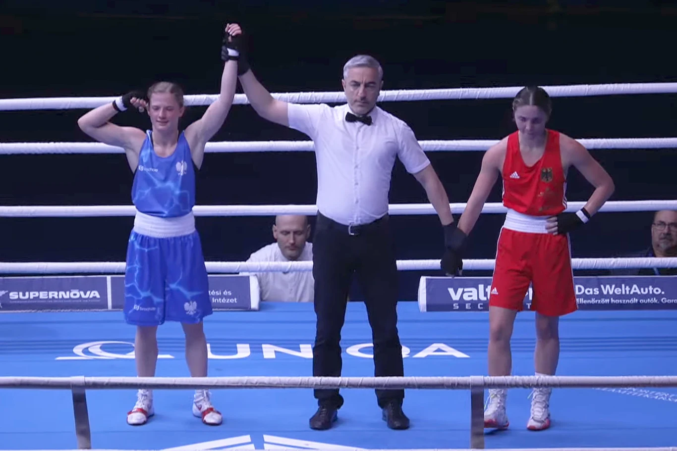 Sędzia podnosi rękę bokserki w niebieskim stroju, ogłaszając jej zwycięstwo w pojedynku. Obok stoi rywalka w czerwonym stroju z opuszczoną głową oraz ręką. Obie zawodniczki znajdują się na ringu bokserskim, widoczne są liny oraz oznaczenia sponsorów na...