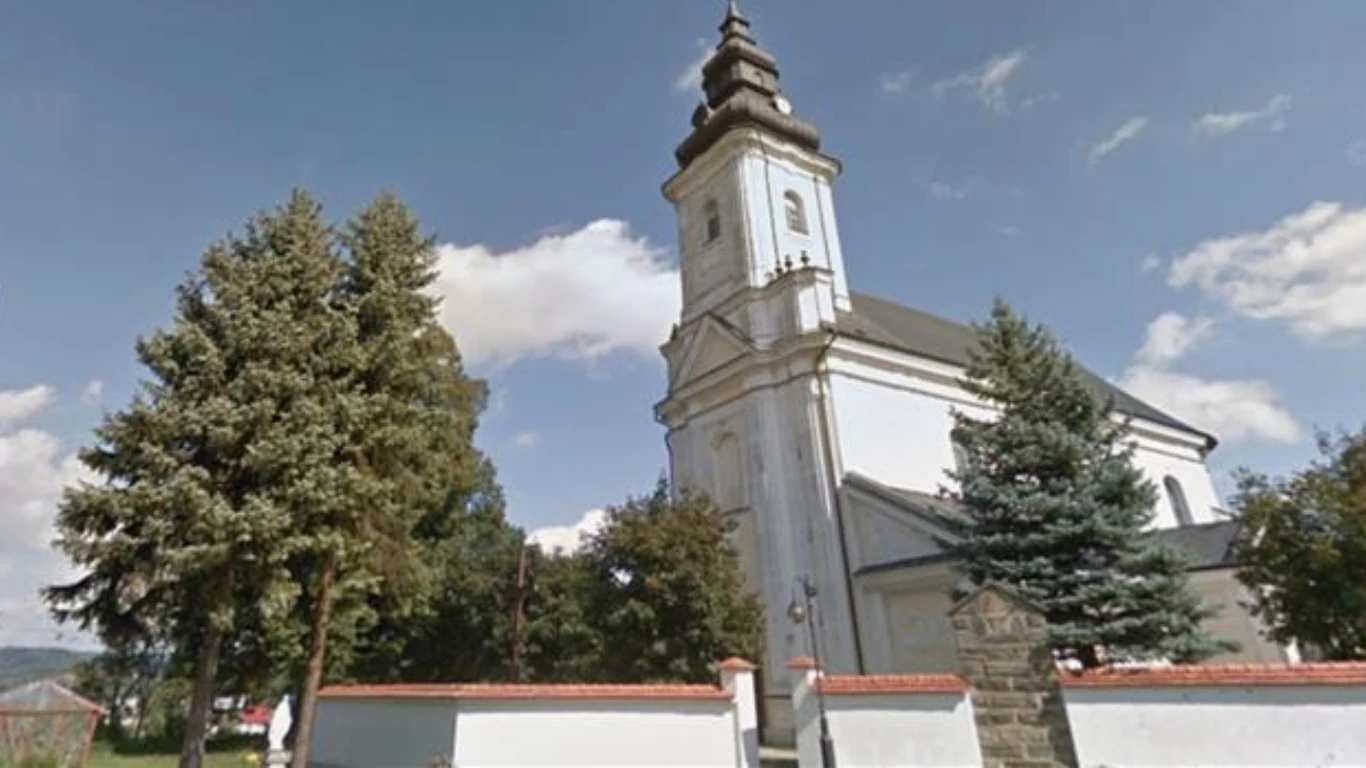 Parafia pw. św. Jakuba Starszego Apostoła w Tuchowie Biała, klasycystyczna świątynia z wysoką wieżą otoczona zielonymi drzewami oraz fragmentem białego ogrodzenia z ceglaną zabudową, na tle niebieskiego nieba z białymi chmurami.