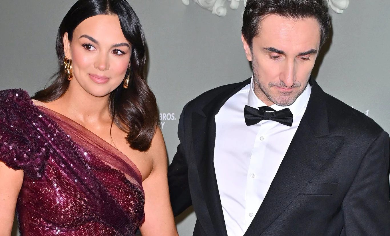 Paulina Krupińska i Sebastian Karpiel-Bułecka Elegancko ubrana para, kobieta w połyskującej bordowej sukni z jednym odkrytym ramieniem oraz mężczyzna w czarnym garniturze i muszce, pozująca razem na tle neutralnej ściany.