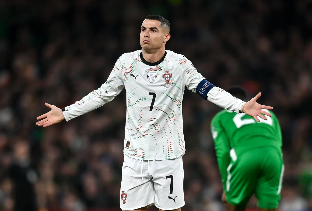 Cristiano Ronaldo Piłkarz w białej koszulce Portugalii z numerem 7 i opaską kapitana w geście rozłożonych rąk, w tle zawodnik w zielonym stroju pochylony do przodu.