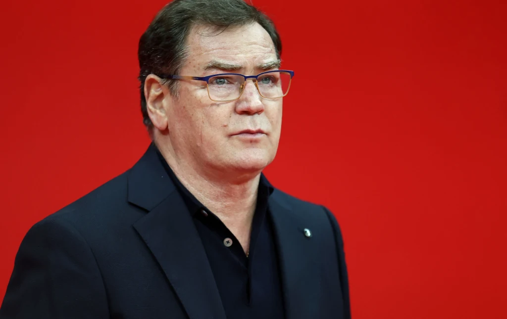 Selekcjoner Jan Urban Mężczyzna w średnim wieku w okularach, ubrany w ciemny garnitur i ciemną koszulę, patrzący w bok, na tle intensywnie czerwonej ściany.