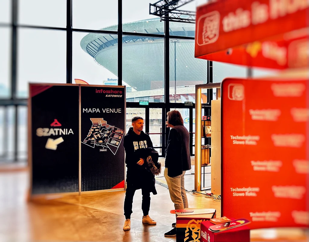 Dwie osoby rozmawiają w katowickim MCK (jedną z nich jest Michał Sadowski z Brand24), podczas konferencji Infoshare 2025, widoczny jest plan wydarzenia oraz oznaczenie szatni. W tle przez duże okna widać charakterystyczny budynek, halę widowiskową Spodek.