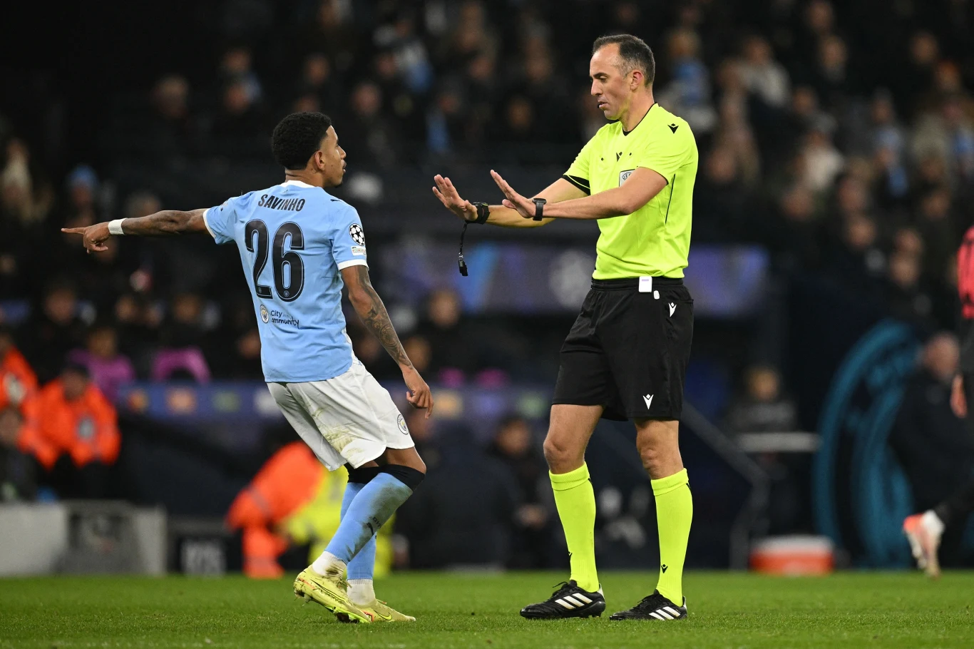 Zawodnik drużyny w niebieskiej koszulce wykonuje dynamiczny ruch podczas dyskusji z sędzią w żółtej koszulce na boisku piłkarskim, w tle widoczni kibice i trybuny.