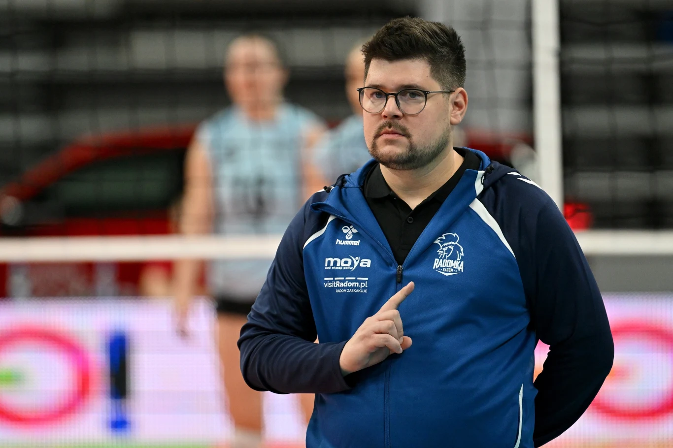 Mężczyzna w sportowej bluzie z logo drużyny, stojący na tle siatkarskiego boiska, w tle rozmazane sylwetki zawodniczek.