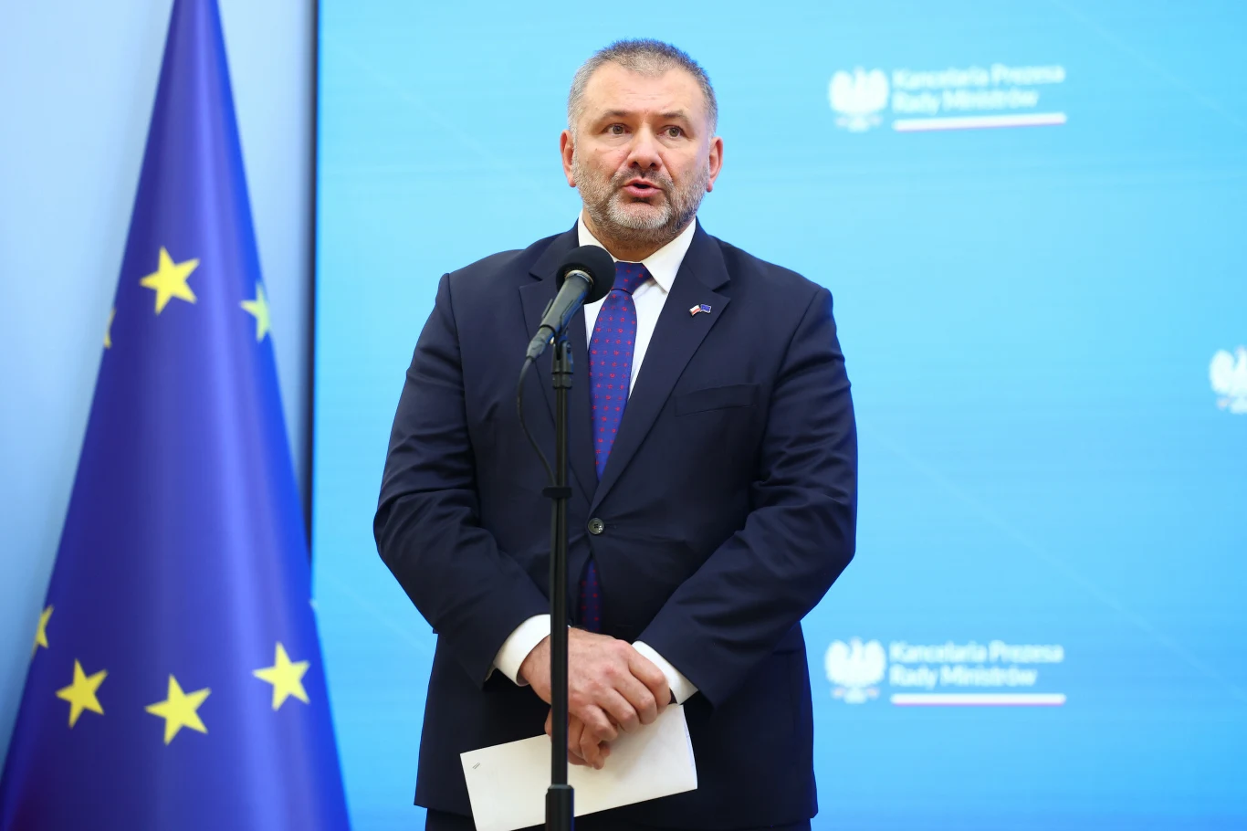 Mężczyzna w garniturze przemawia do mikrofonu na tle niebieskiej ścianki z logotypami i flagi Unii Europejskiej, trzyma w rękach kartkę papieru.