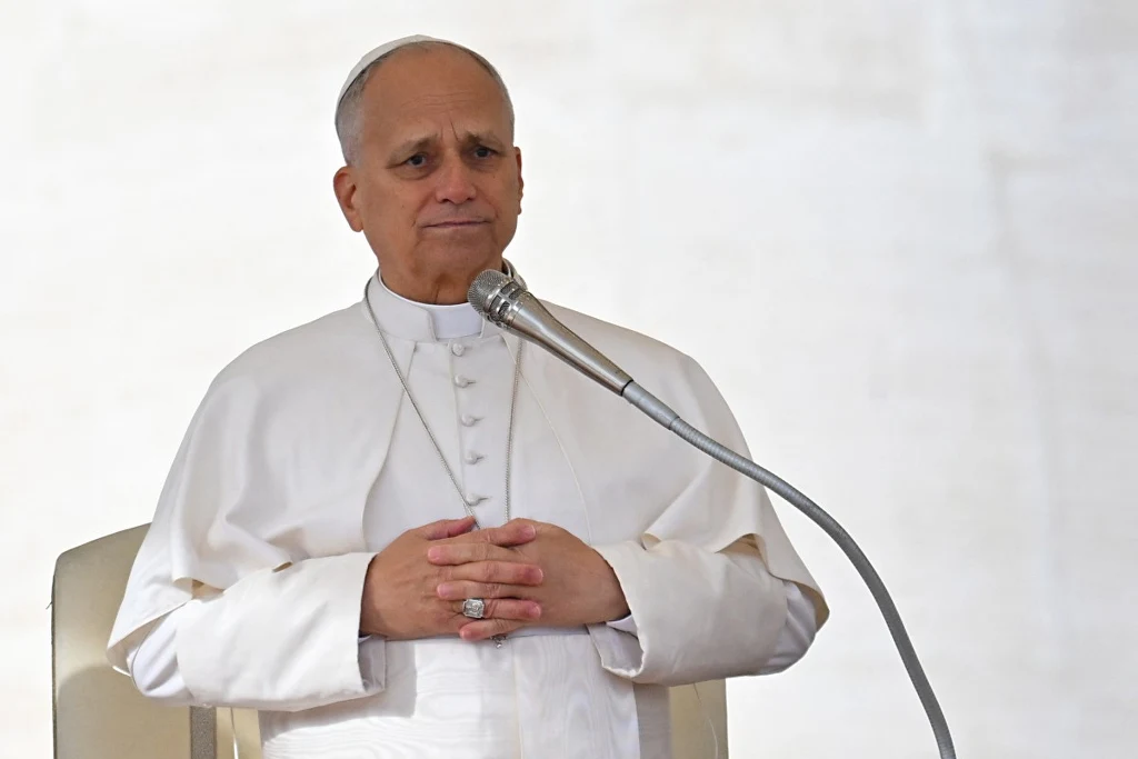 Papież Leon XIV Starszy duchowny ubrany w biały strój papieski siedzi na jasnym tle, trzyma dłonie splecione przed sobą, przed nim mikrofon na stojaku.