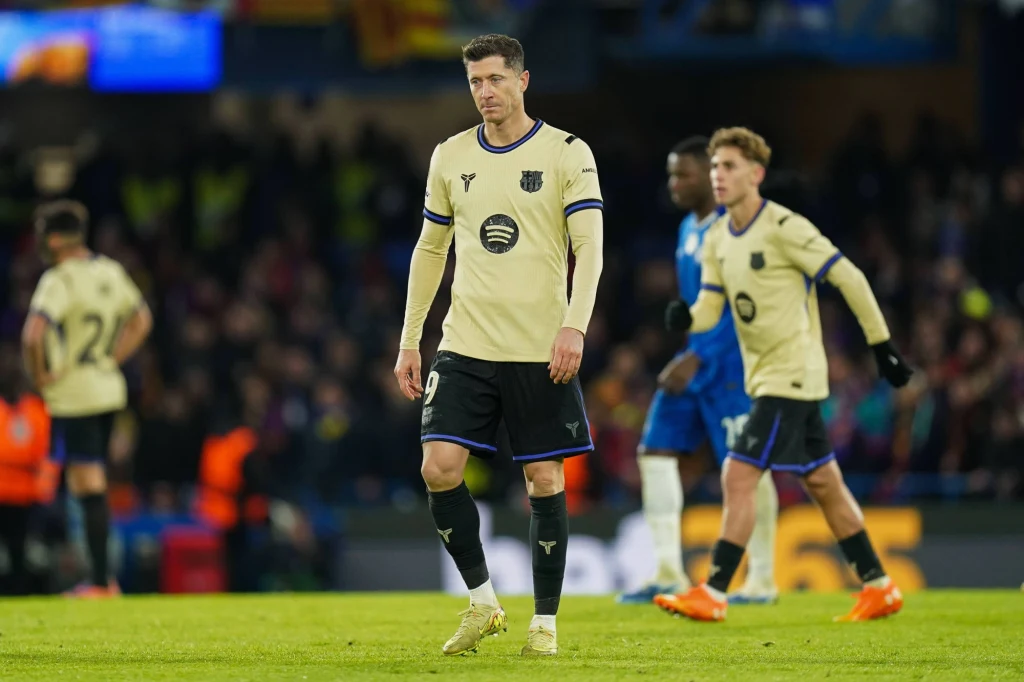 Robert Lewandowski w meczu Ligi Mistrzów przeciwko Chelsea Zawodnik drużyny piłkarskiej w jasnym stroju schodzi po murawie z wyrazem niezadowolenia na twarzy. W tle zawodnicy przeciwnej drużyny oraz kibice na trybunach.