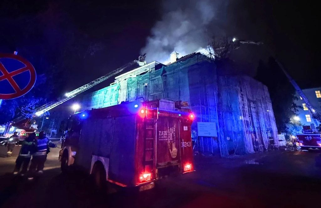 Strażacy gaszą pożar budynku otoczonego rusztowaniem, wokół rozstawione samochody strażackie, z dachu unosi się dym, a nocne oświetlenie podkreśla intensywność akcji.