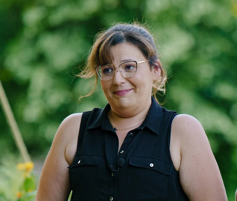 Kobieta w okularach, uśmiechnięta, ubrana w czarną koszulę bez rękawów, stoi na tle rozmytego, zielonego ogrodu w słoneczny dzień.