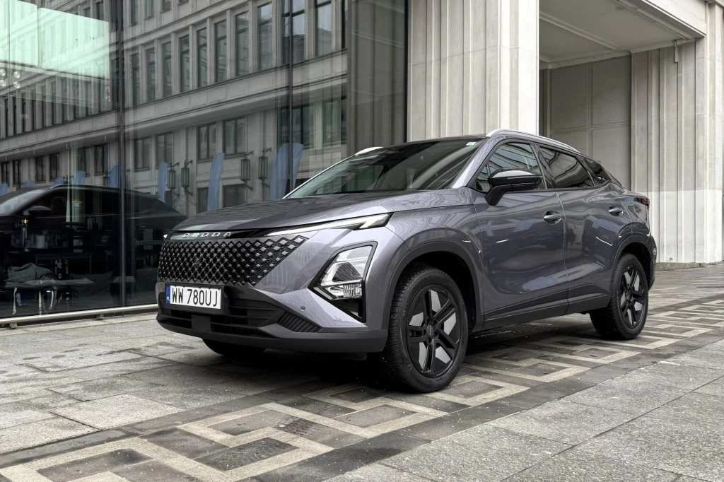 Nowoczesny SUV w grafitowym kolorze zaparkowany przed szklano-betonowym budynkiem, jego reflektory oraz ostra linia nadwozia podkreślają dynamiczny charakter pojazdu, w tle odbicie miasta w dużych oknach