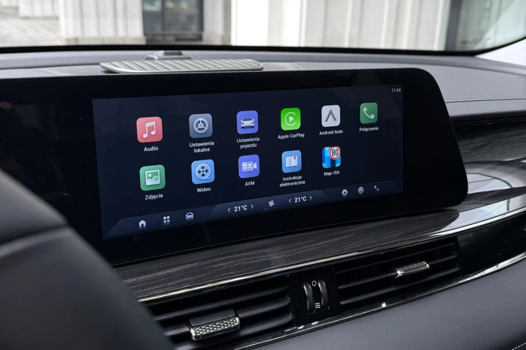 System multimedialny samochodu z dużym dotykowym ekranem wyświetlającym menu główne z ikonami aplikacji, takich jak audio, ustawienia pojazdu, nawigacja, klimatyzacja oraz integracje z Apple CarPlay i Android Auto.