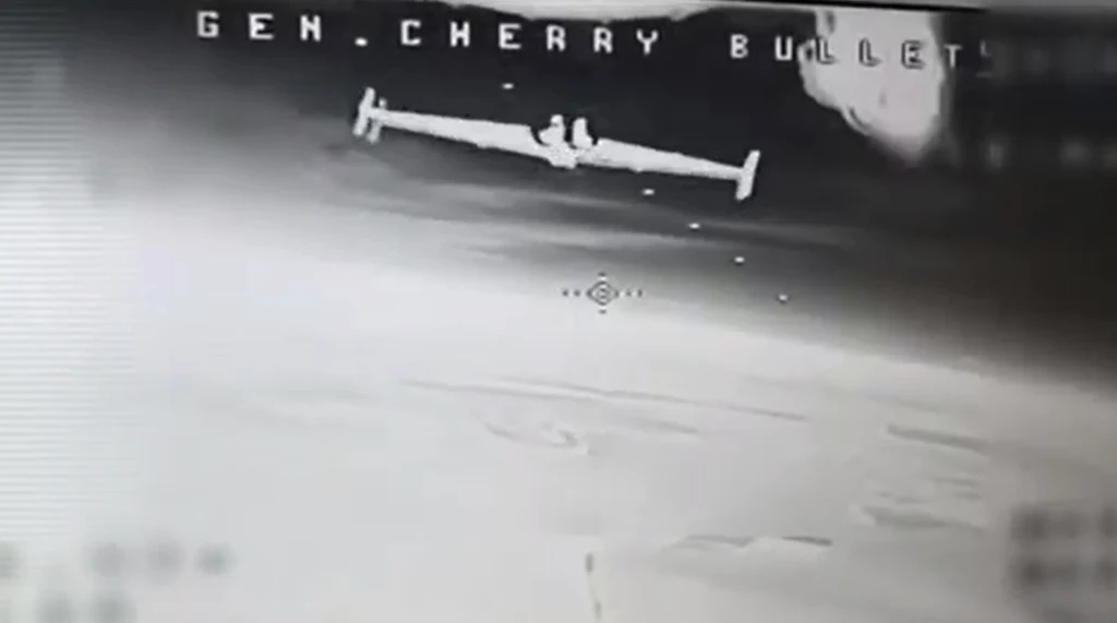 Ujęcie z monitora ukazujące czarno-biały obraz drona wojskowego lecącego nad rozległym obszarem ziemi, w tle cyfrowe oznaczenia i celownik.