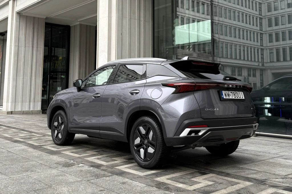 Nowoczesny szary SUV zaparkowany przy krawężniku przed szklaną witryną i wysokim budynkiem miejskim, odbicia w szybach podkreślają urbanistyczny charakter otoczenia.