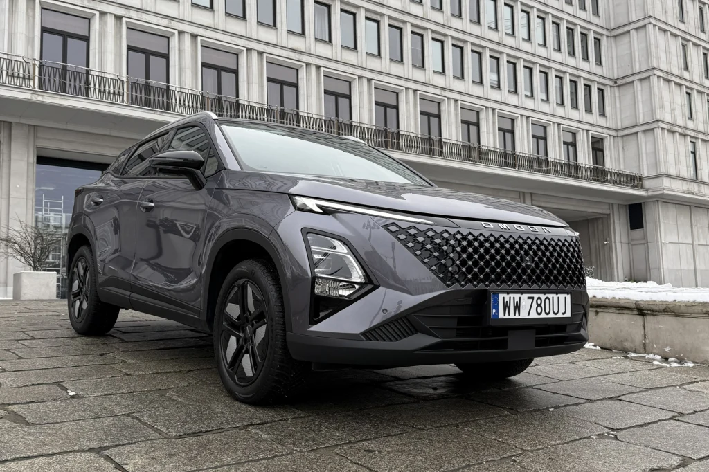 Nowoczesny szary SUV zaparkowany przed betonowym biurowcem z wieloma oknami, widoczny z niskiej perspektywy. Samochód posiada czarne felgi i wyróżnia się charakterystycznym grillem oraz polską rejestracją.