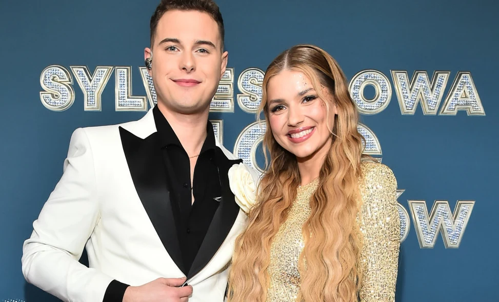 Adam Zdrójkowski i Julia Żugaj. Młody mężczyzna w eleganckim, białym garniturze z czarną koszulą oraz młoda kobieta z długimi, kręconymi blond włosami w błyszczącej sukience stoją uśmiechnięci razem na tle niebieskiej ściany z dużym napisem.