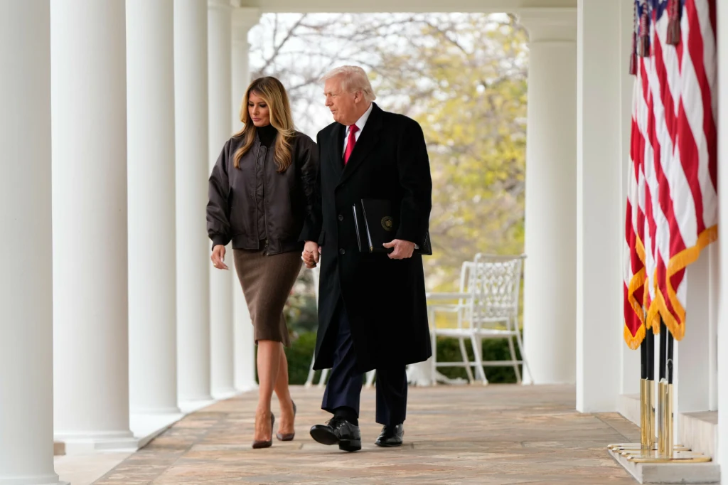 Stylizacja Melanii Trump była przemyślana w każdym calu Mężczyzna w ciemnym płaszczu i czerwoną krawatą spaceruje trzymając za rękę kobietę w eleganckiej spódnicy i kurtce, oboje idą alejką z kolumnami, w tle widoczna amerykańska flaga oraz ławka ogrodowa.
