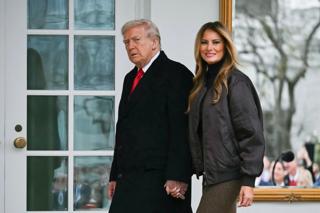 Melania tym razem wybrała mniej wystawną stylizację Para w eleganckich ubraniach trzymająca się za ręce, mężczyzna w czarnym płaszczu z czerwonym krawatem, kobieta w ciemnej kurtce z długimi rozpuszczonymi włosami, w tle szklane drzwi i odbicie osób na zewnątrz.