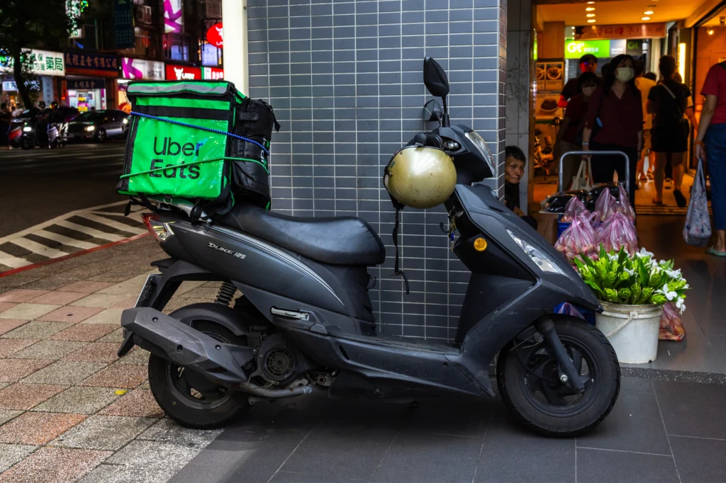 Dwusuwowe skutery powinny zniknąć z dróg? Zdj. ilustracyjne Czarny skuter z torbą kurierską Uber Eats zaparkowany przy szarej ścianie obok wejścia do sklepu, w tle kilka osób robiących zakupy i ustawione przy wejściu kwiaty.