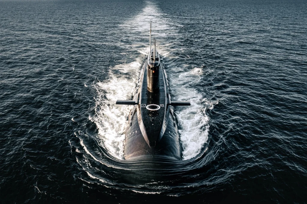Okazały okręt podwodny płynący po powierzchni otwartego morza, widziany z góry w centrum kadru z wyraźnym śladem piany za rufą. Wokół rozciąga się szeroka przestrzeń wodna.