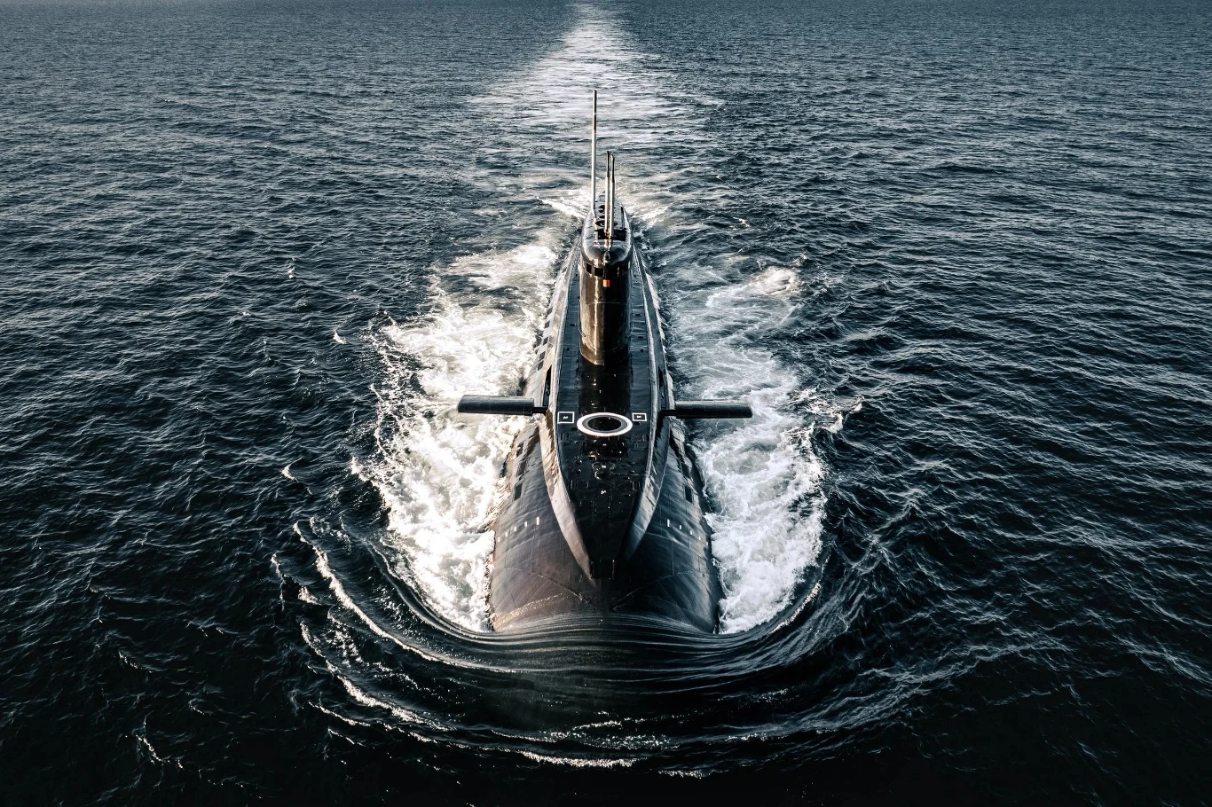Okazały okręt podwodny płynący po powierzchni otwartego morza, widziany z góry w centrum kadru z wyraźnym śladem piany za rufą. Wokół rozciąga się szeroka przestrzeń wodna.