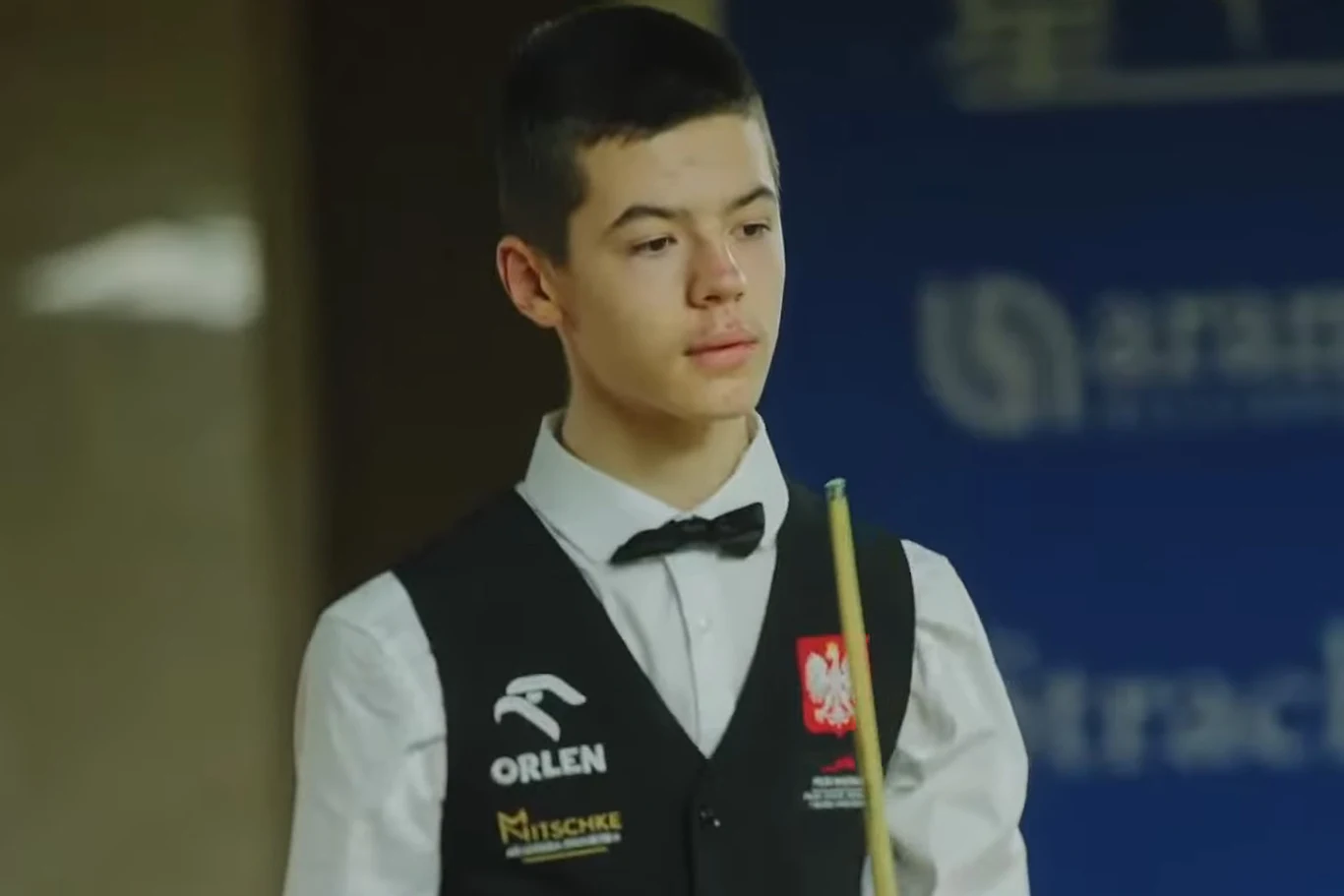 Młody zawodnik bilardowy w stroju turniejowym z kamizelką, muszką i kijem bilardowym, skupiony podczas rozgrywki, na tle niebieskiego banera z logo sponsora.