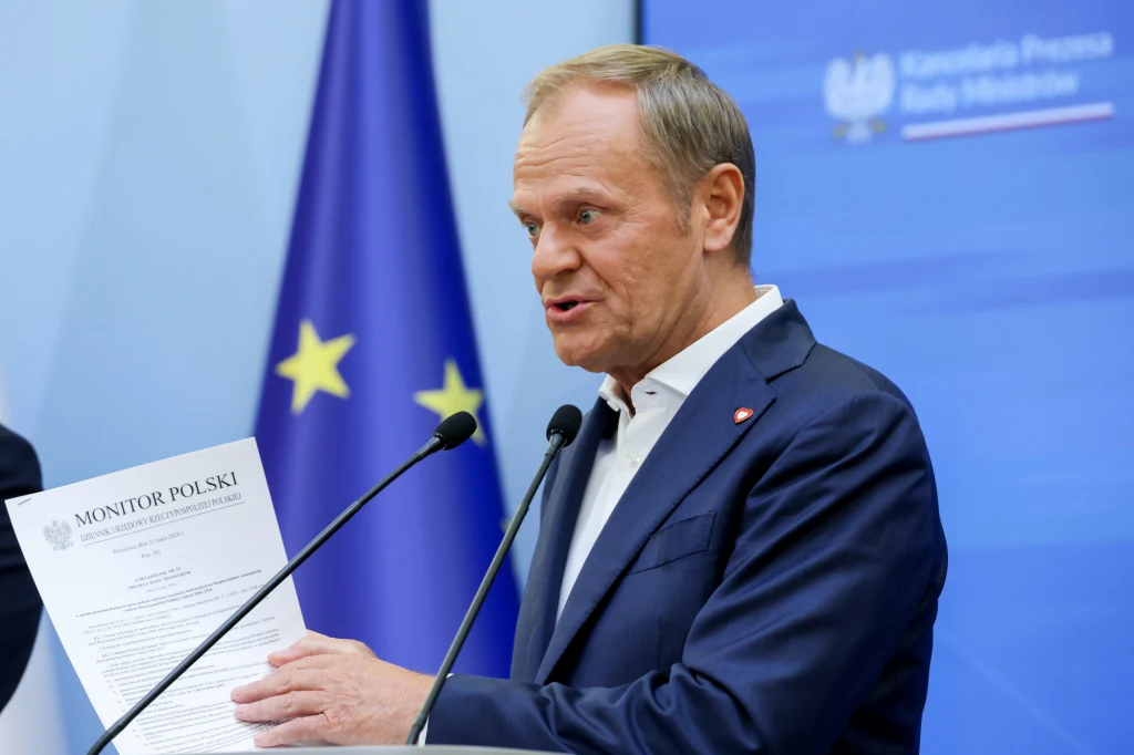 Komisję ds. badania wpływów rosyjskich i białoruskich w maju 2024 roku powołał premier Donald Tusk Mężczyzna w garniturze przemawia do mikrofonów, trzymając w ręku dokument Monitor Polski. W tle widoczne flagi Unii Europejskiej oraz tablica z napisem Kancelaria Prezesa Rady Ministrów.