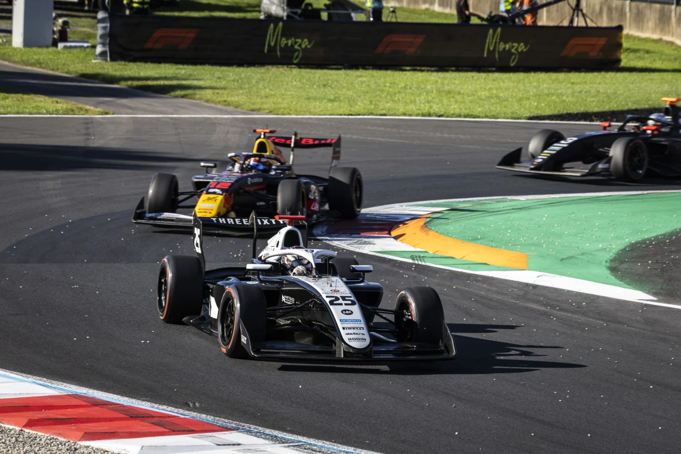 Trzy bolidy Formuły 2 rywalizują na zakręcie toru wyścigowego Monza, pośród zielonych traw i asfaltu, z reklamami charakterystycznymi dla toru w tle.