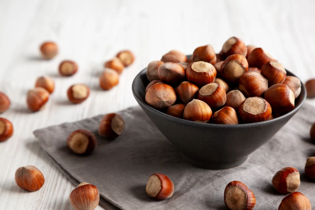 Jedz garść laskowców dziennie. Obniżysz cukier, cholesterol i poprawisz pamięć. Czarna ceramiczna miska wypełniona orzechami laskowymi ustawiona na szarej serwetce, wokół miski rozsypane są pojedyncze orzechy na jasnym drewnianym blacie.