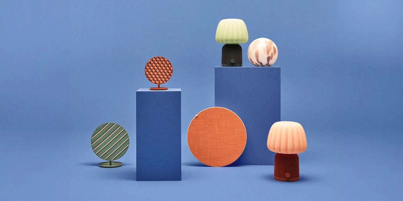 Ikea KULGLASS i SOLSKYDD. Nowe głośniki Bluetooth, które są kolorowe. Kolorowe, designerskie lampki stołowe oraz okrągłe przedmioty dekoracyjne ustawione na niebieskim tle i geometrycznych podestach, przedmioty utrzymane w pastelowej i stonowanej kolorystyce.