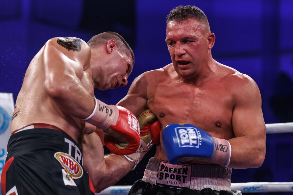 Mateusz Tryc (z lewej) na zdjęciu z kwietniowej walki, gdy rywalem był Niemiec Nick Hannig Dwóch bokserów w trakcie starcia na ringu, jeden z zawodników wyprowadza cios na korpus przeciwnika, obydwaj są spoceni i skoncentrowani, wokół widać atmosferę wydarzenia sportowego.