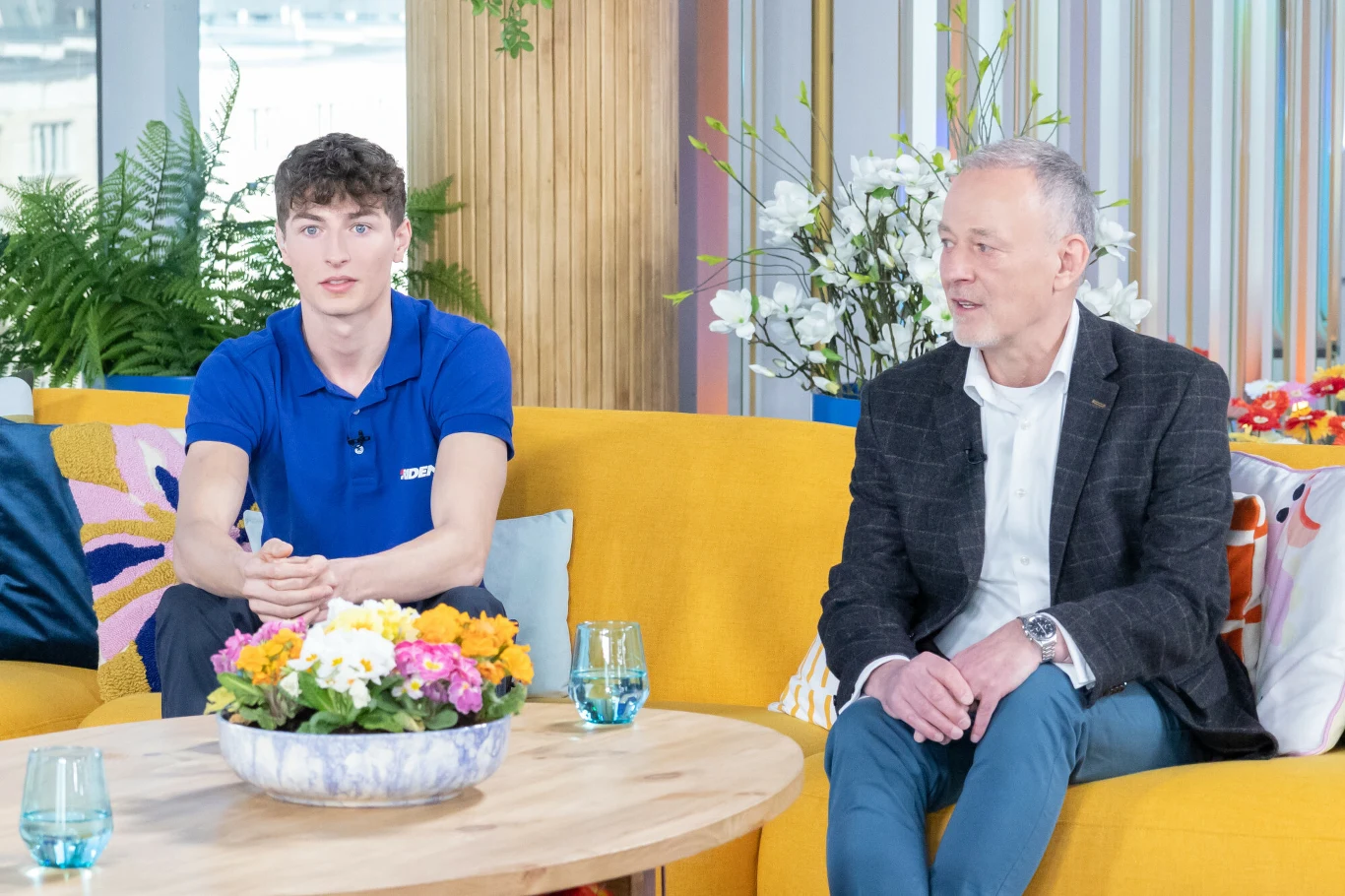 Dwóch mężczyzn siedzących na żółtej sofie w nowocześnie urządzonym wnętrzu, jeden młodszy w niebieskiej koszulce, drugi starszy w marynarce, na stole przed nimi bukiet kwiatów i szklanki z wodą, w tle rośliny i dekoracje.