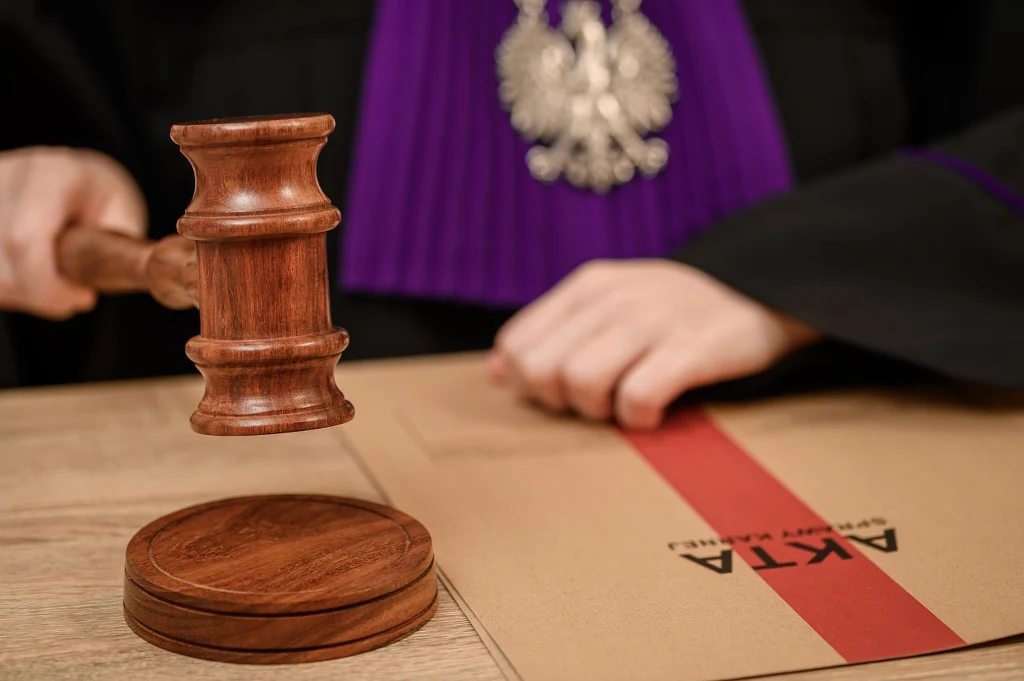 Młotek sędziowski umieszczony na drewnianym blacie, dłonie osoby w togach sędziowskich z widocznym godłem Polski oraz duża koperta z napisem 'AKTA' przewiązana czerwoną taśmą