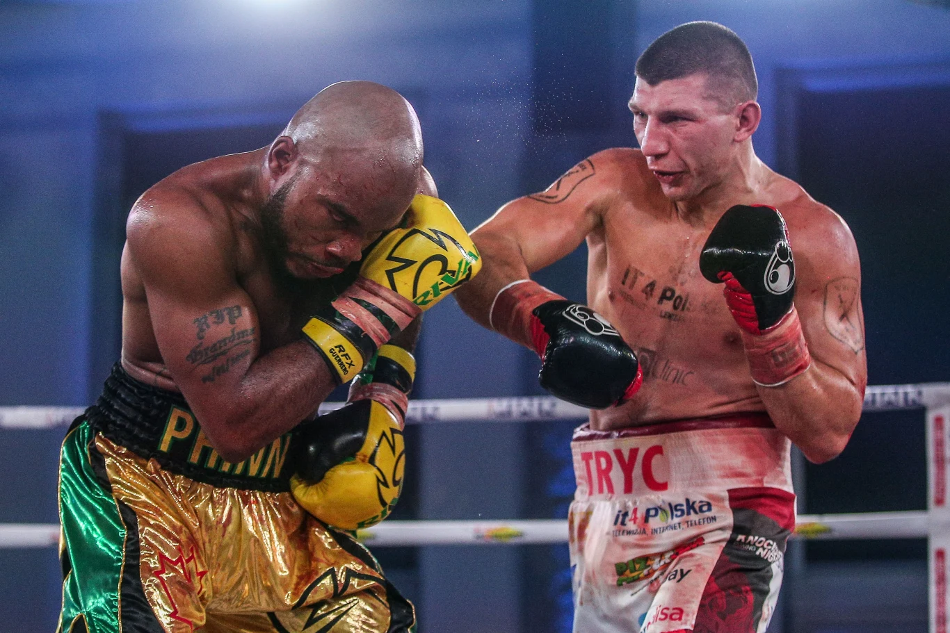 Dwóch bokserów walczących w ringu, jeden ubrany w złote i czarne spodenki jest w defensywie, osłania się przed ciosem, drugi bokser z widocznymi tatuażami i zakrwawioną twarzą zadaje uderzenie prostym prawym.