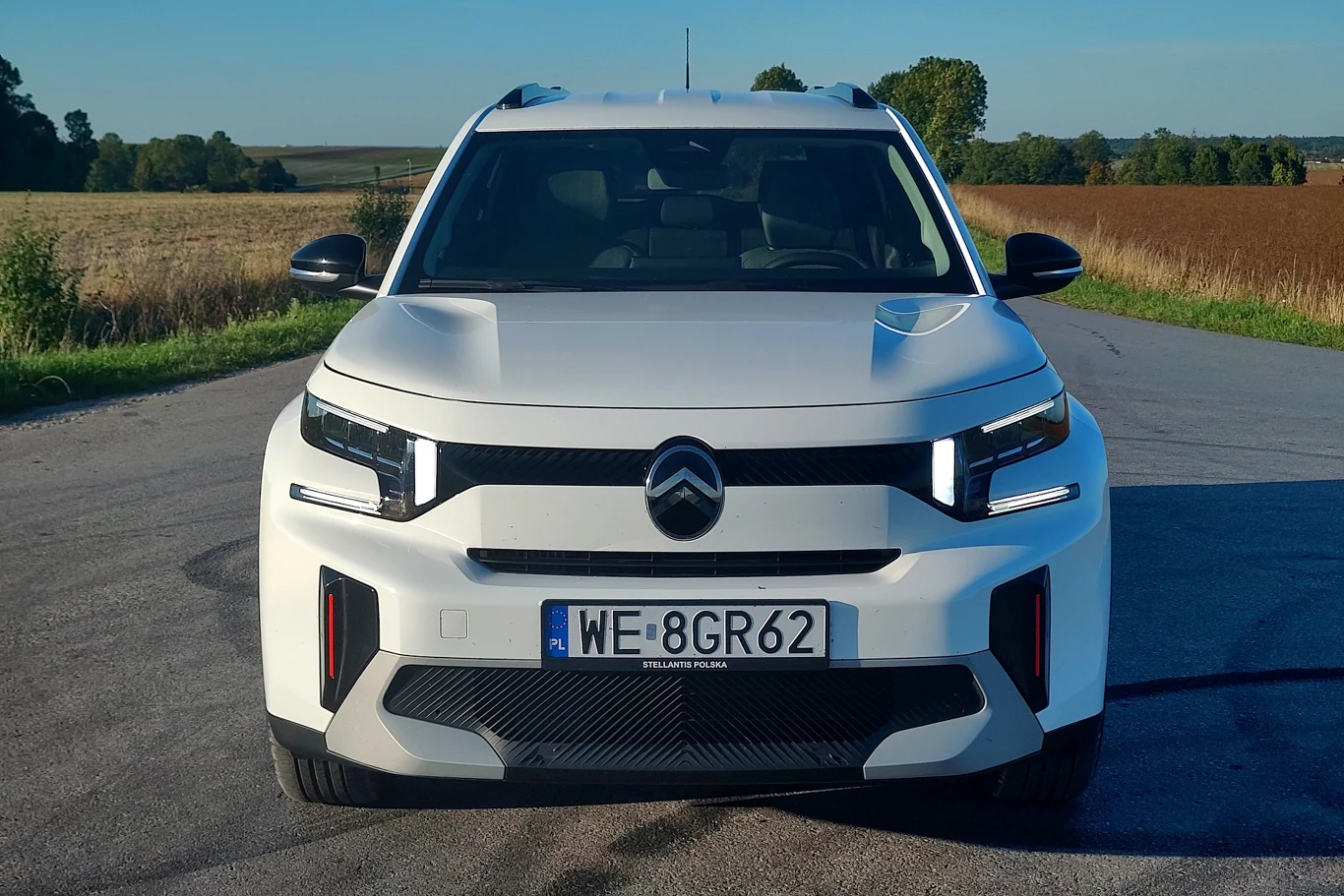 Biały, nowoczesny samochód Citroën C3 Aircross widziany z przodu, stoi zaparkowany na asfaltowej drodze, otoczonej polami i drzewami w tle.