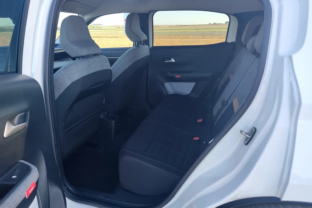Na tylnej kanapie miejsca jest w bród, ale dotyczy to wersji pięcioosobowej. W siedmioosobowej kanapa na stałe jest przesunięta do przodu o 6,5 cm Wnętrze tylnej części samochodu osobowego z widokiem na dwa tylne siedzenia i fragment przednich foteli, wykończenie foteli w odcieniach szarości i czerni, szerokie okno ukazujące krajobraz pól w tle.